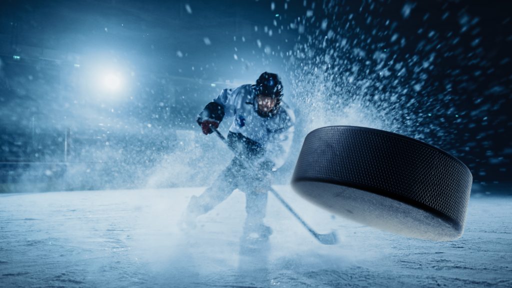 "Ishockeyspelare i vit tröja skjuter pucken hårt på isen medan snö och is sprutar upp omkring honom."