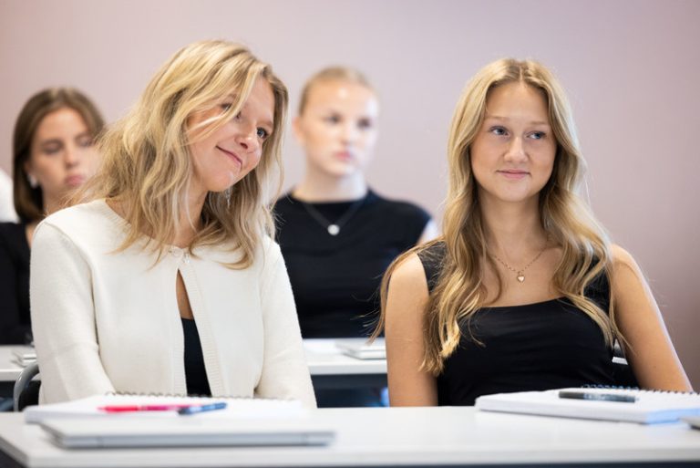 Två unga kvinnor sitter bredvid varandra vid ett skrivbord i ett klassrum, båda ler svagt och tittar åt samma håll. Framför dem ligger anteckningsblock och pennor. I bakgrunden syns andra studenter i oskärpa.