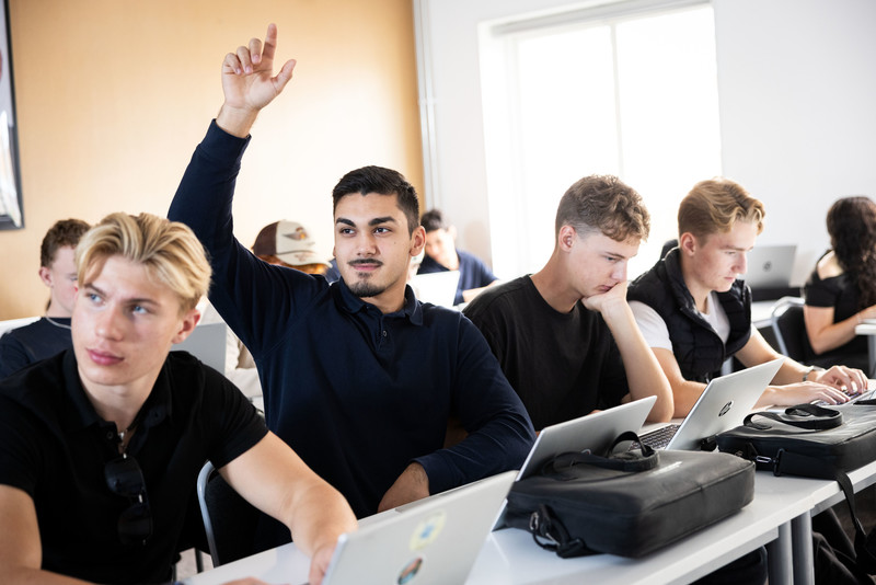 En grupp gymnasieelever sitter vid sina datorer i ett klassrum. En av eleverna räcker upp handen för att ställa en fråga.