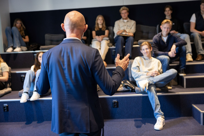 En gymnasieklass sitter ner och tittar och lyssnar på lärare som står upp och undervisar. Bild tagen bakifrån på läraren med elever i fokus.