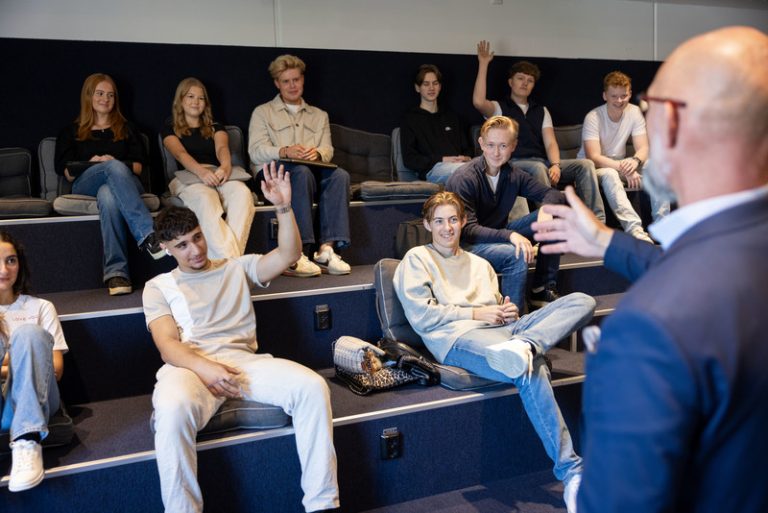 Lärares ryggtavla i fokus, som pratar till elever som sitter på en gradäng och räcker upp händerna, samt ser intresserad in i kameran eller på läraren.