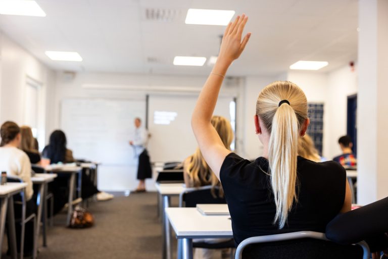 Elev som räcker upp handen i fokus i ett vitt klassrum. Man ser andra elever suddigt och även lärare framför.