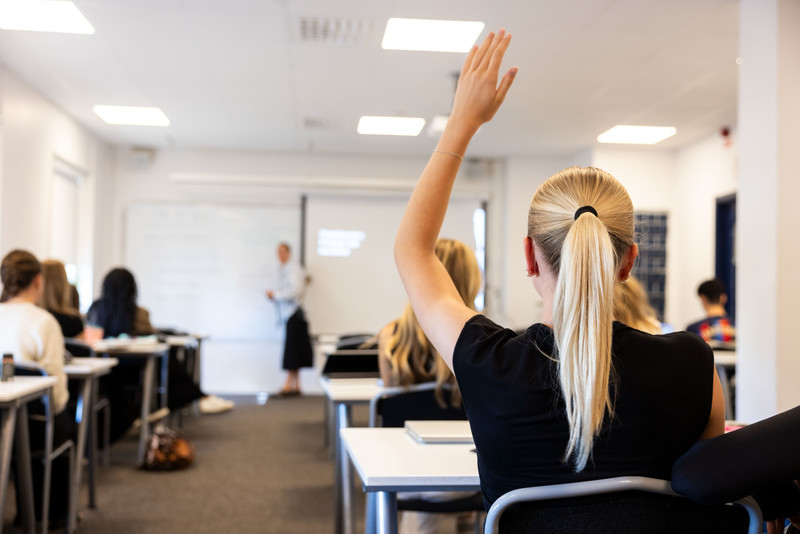 Lektion, bild tagen bakifrån på en hel klass. Fokus på elev som sitter och räcker upp handen inför läraren.
