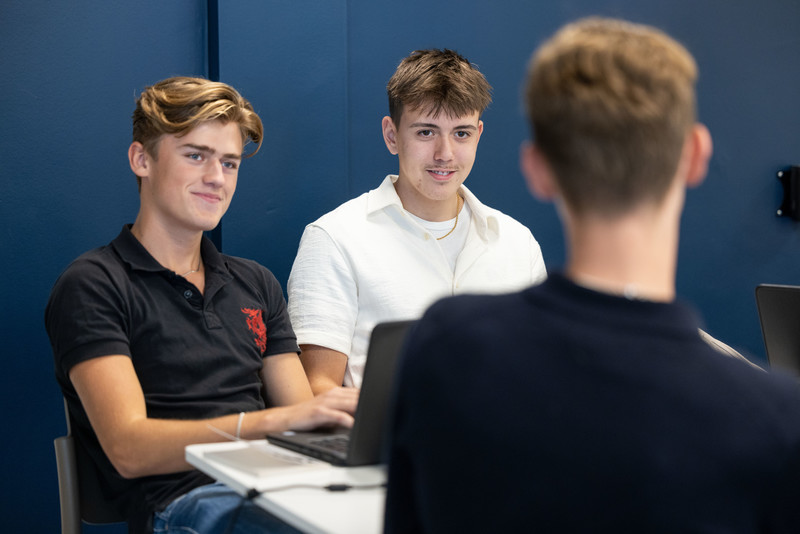 Tre manliga gymnasieelever sitter tillsammans med sina datorer och ser glada ut under en rast.