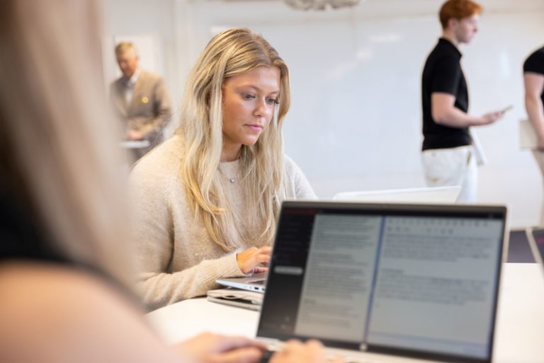 En ung tjej är i fokus och sitter och arbetar i ett klassrum. Man kan också urskilja en datorskärm.