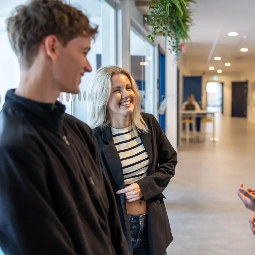 Närbild på en tjej och en kille som står i en korridor på affärsgymnasiet, de har en konversation med en tredje elev som inte syns i bild.
