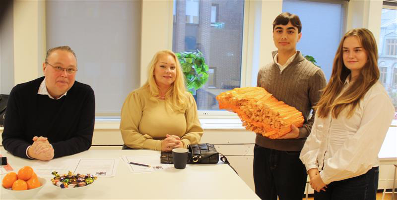 Fyra personer sitter och står vid ett bord i ett klassrum. Två elever håller i en orange trämodell, medan två vuxna sitter vid bordet med papper, kaffe och godis framför sig.