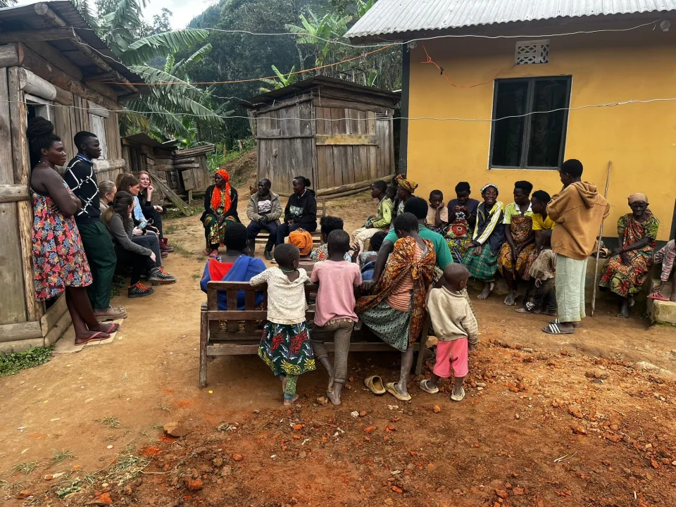 En grupp människor, både vuxna och barn, sitter samlade utomhus i en bymiljö mellan enkla trähus och ett gult hus. Några sitter på bänkar medan andra står runt omkring, som i ett möte eller samtal i samhället.