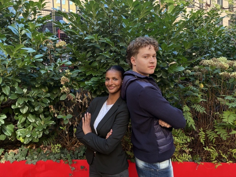 En ung kvinna och en ung man står rygg mot rygg med armarna i kors framför en grön buske i en stadsmiljö. Kvinnan ler och bär svart kavaj och vit t-shirt. Mannen har lockigt hår, mörkblå tröja och jeans. I förgrunden syns en röd kant och i bakgrunden en ljus byggnad.