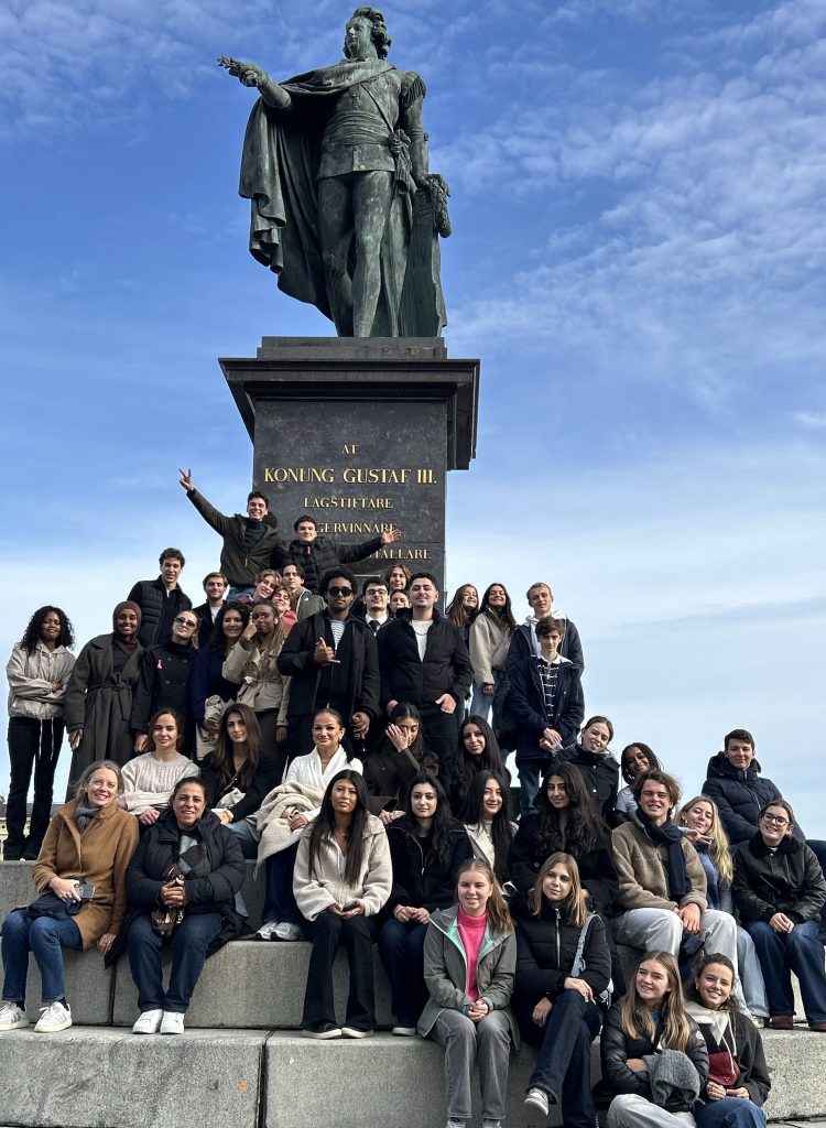 Stor gruppbild med ett femtiotal ungdomar och några vuxna som sitter och står på trappstegen kring en staty under en blå himmel. Statyn i grön brons föreställer en man i mantel med utsträckt arm, och på sockeln står med guldbokstäver ”Åt Konung Gustaf III”. Några i gruppen gör segertecken och poserar glatt.