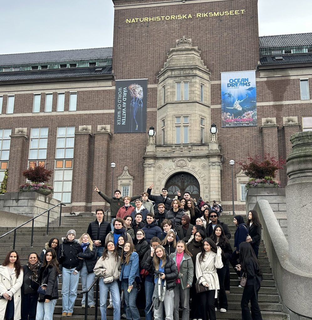 En stor grupp ungdomar och några vuxna står samlade på en bred stentrappa framför en tegelbyggnad med texten ”Naturhistoriska riksmuseet” högst upp. På fasaden hänger banderoller för utställningen ”Värld av vatten” och filmen ”Ocean Dreams”. Några i gruppen sträcker upp armarna och gör segertecken.