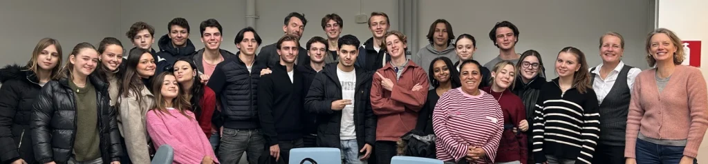Gruppfoto i ett klassrum med ett trettiotal personer, de flesta tonåringar, som står tätt tillsammans och ler mot kameran. Många bär vinterjackor och tröjor. Några gör skämtsamma gester, en pekar på sig själv och en annan putar med läpparna. Längst till höger står två vuxna kvinnor, en i rosa kofta och en i väst och vit skjorta.